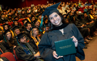 a graduate smiles as she holds up her diploma cover while standing in front of her class of graduates
