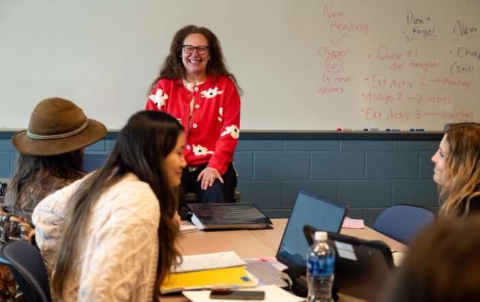 an instructor at the front of a class smiles as do a few students doing classwork at tables in front of her