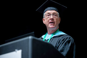 Jon Longest wearing graduation regalia and speaking at the podium