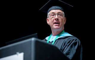 Jon Longest wearing graduation regalia and speaking at the podium