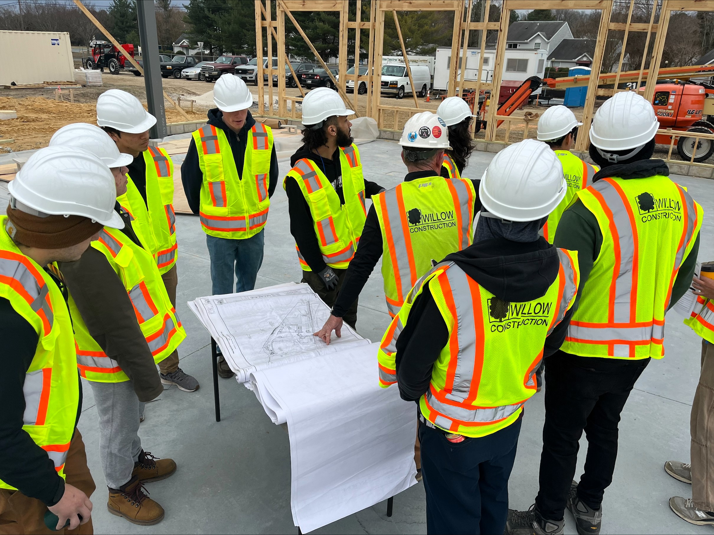 On person with his hand on a blueprint at a construction site motions to everyone else, wearing safety vests and hard hats, like Project Management for Construction