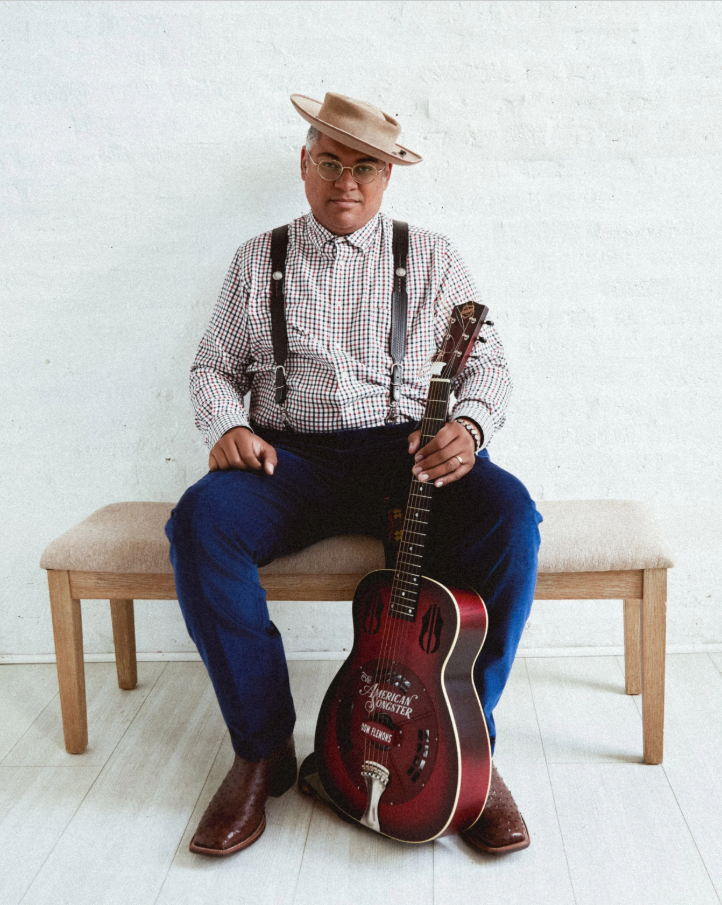 Dom Flemons portrait, sitting on a bench, holding a guitar